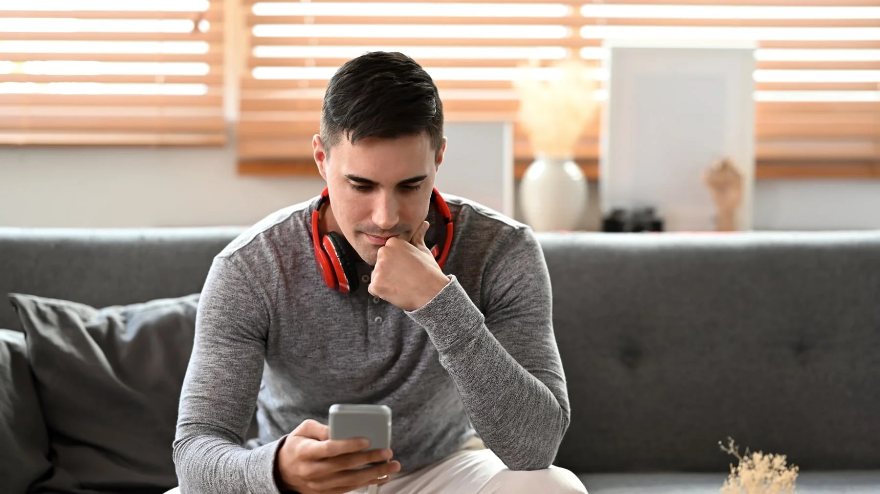 Homem jovem pensativo olhando o celular no sofá, com expressão de dúvida.