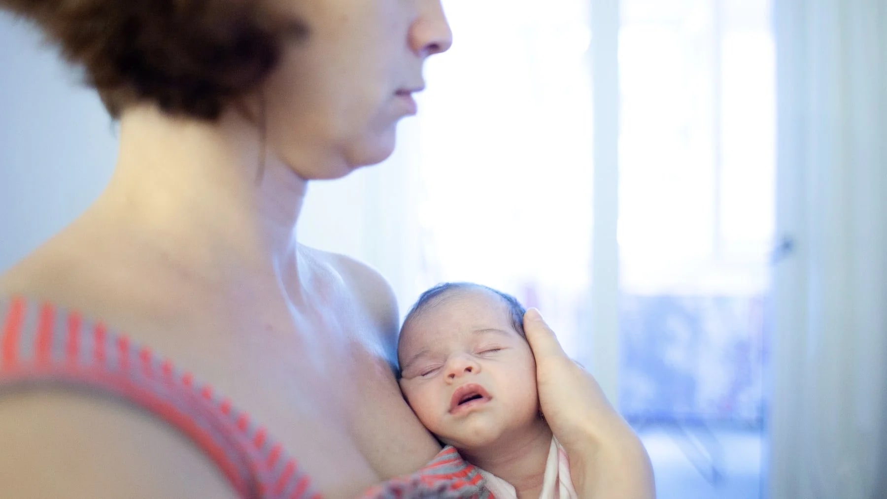 Mãe segurando o bebê recém-nascido junto à janela, com expressão terna e cansada.