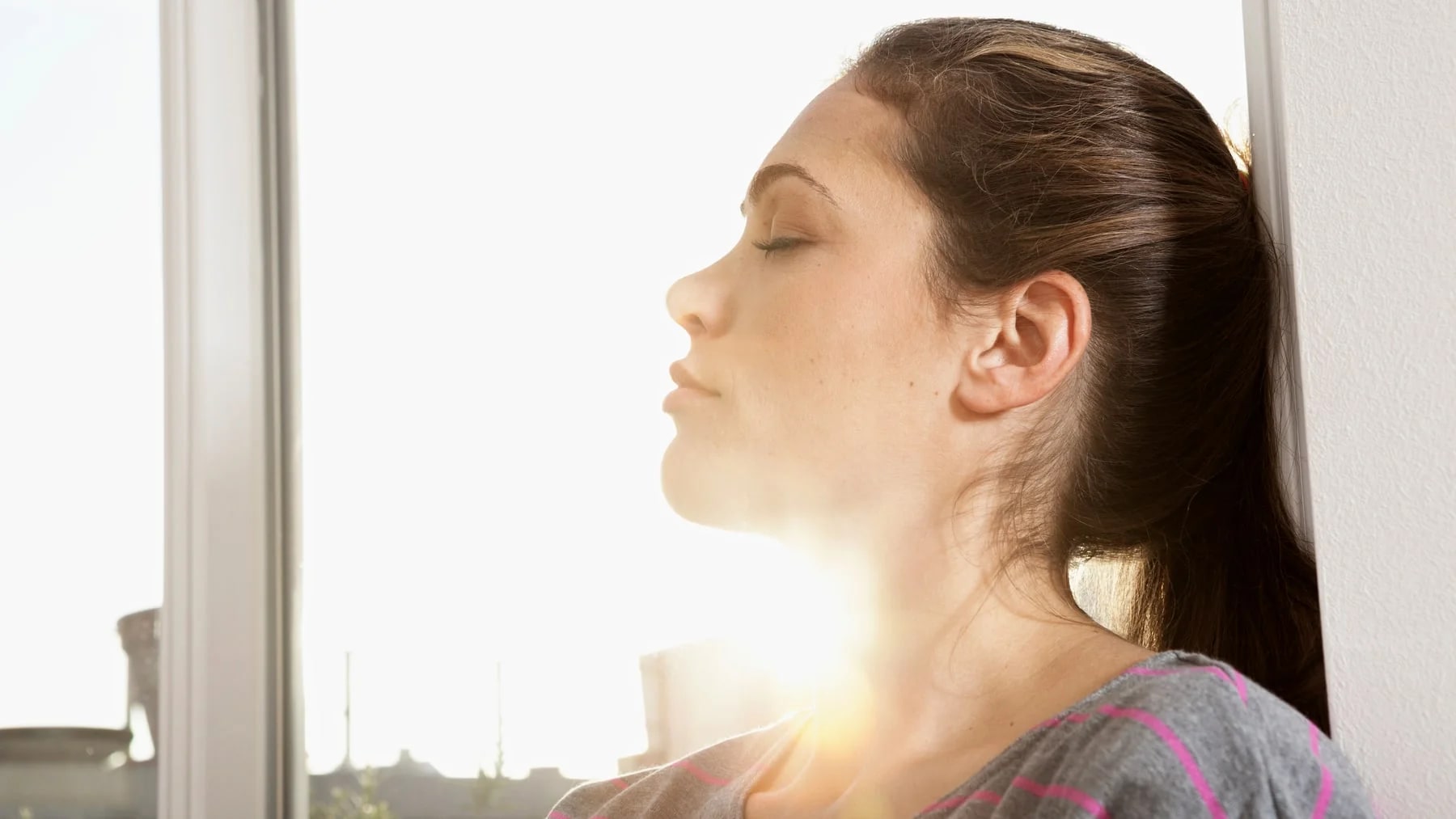 Mulher de olhos fechados recebendo a luz do sol junto à janela, em um momento de calma e respiração.