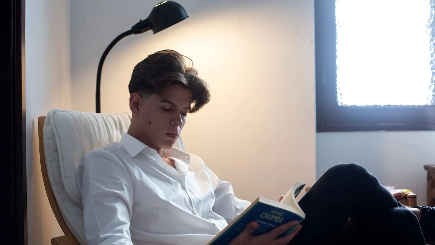 Homem jovem lendo um livro com calma em casa, sob a luz de um abajur.