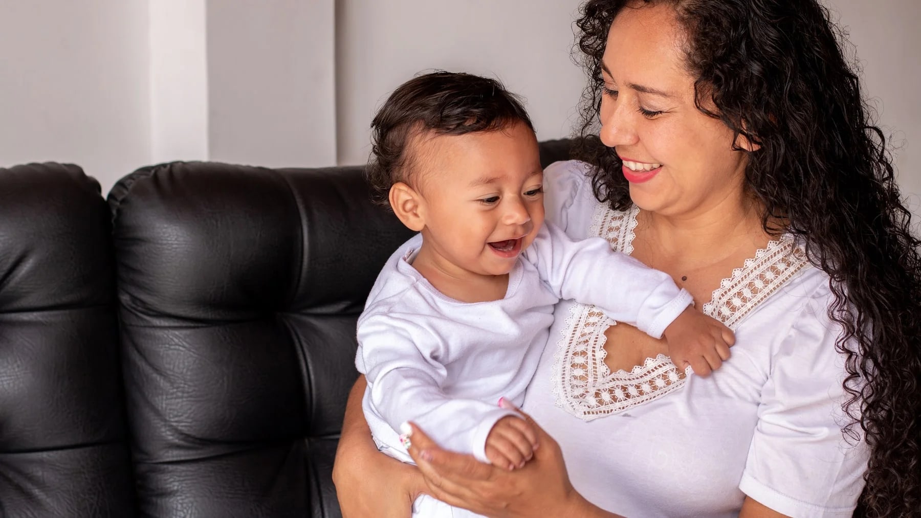 Mãe com o bebê no colo, os dois sorrindo num momento cotidiano em casa.