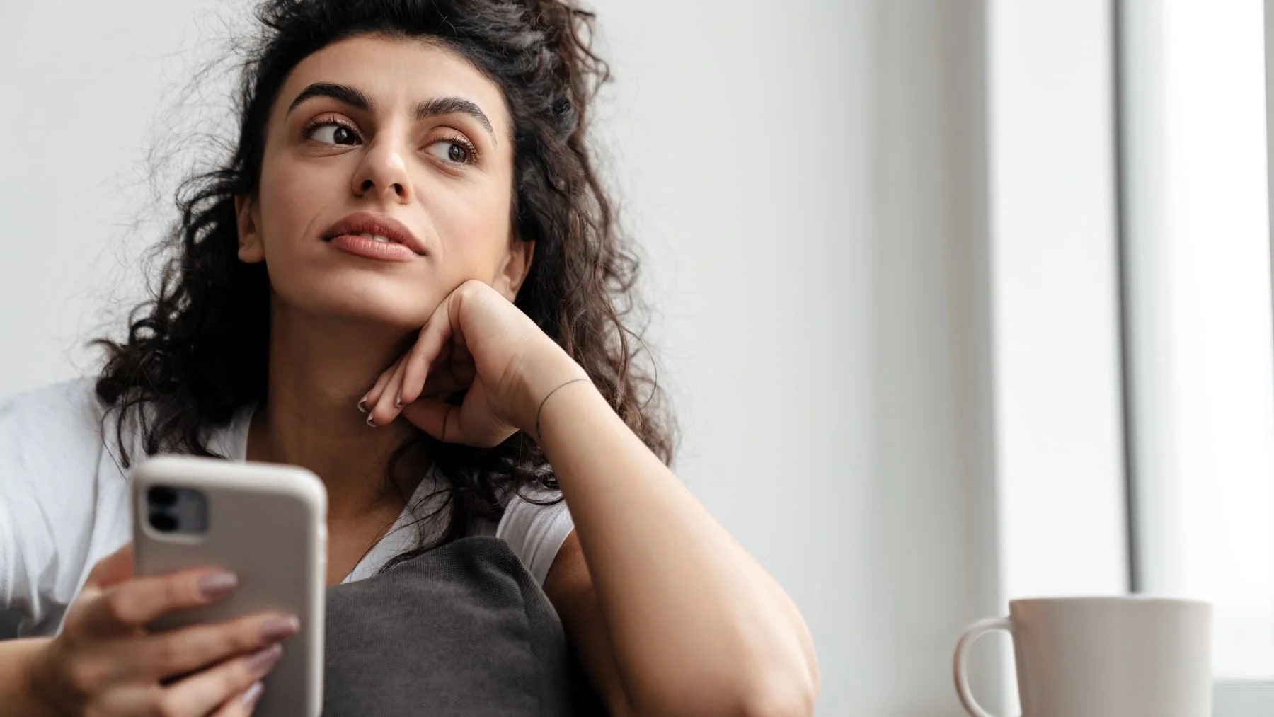 Mulher pensativa em casa com o celular na mão e o olhar levantado, como quem tenta lembrar de algo.