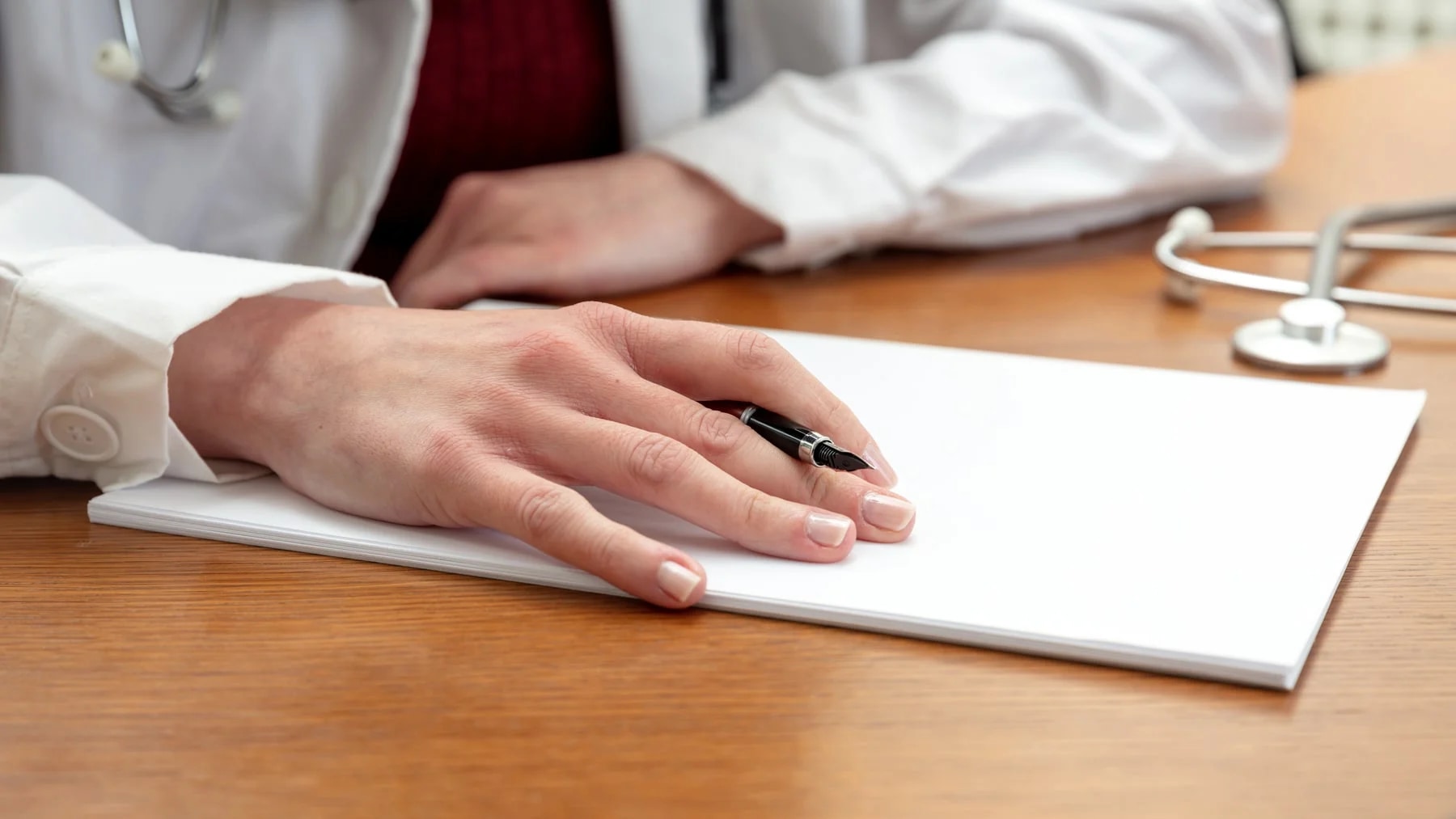 Mão de médica com caneta sobre o papel na mesa do consultório, pronta para anotar.