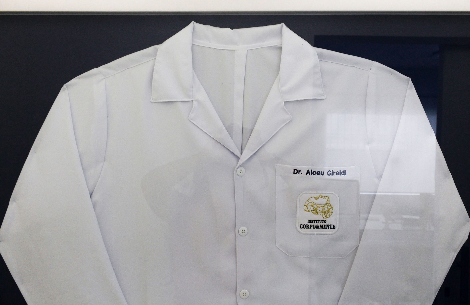 Jaleco original do Dr. Alceu Giraldi, emoldurado na clínica, com nome bordado e logotipo do antigo Instituto Corpo & Mente
